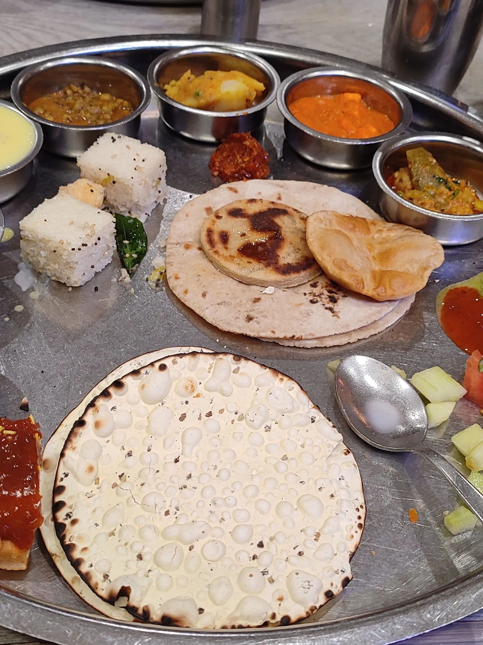 Gujarati vegetarian thali on a steel plate: dhokla, rotli, puri, a large papad and vegetables in small steel bowls